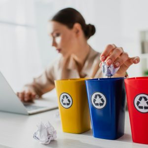selective focus of businesswoman throwing crumpled papers into recycling buckets while working on